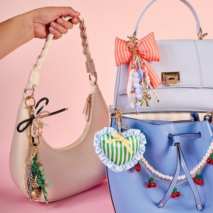 A hand holds a cream handbag with decorative charms. Nearby are blue handbags embellished with bows, beads, pearls, and the-darling-effect’s B Bubble Letter Keychain Bag Charm—personalized—against a pink background.