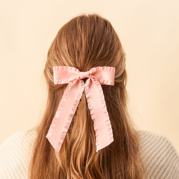 A person with long, wavy light brown hair wears The Darling Effect's Blush Ruffle Hair Bow at the back of their head. Set against a soft beige background, they pair the elegant accessory with a cream sweater.