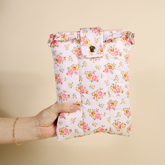 A hand holds the-darling-effect's Endless Daydream Cream Book Cover, a cute, puffy polyester cover with a pink and orange floral pattern and buttoned flap, set against a plain light beige background.