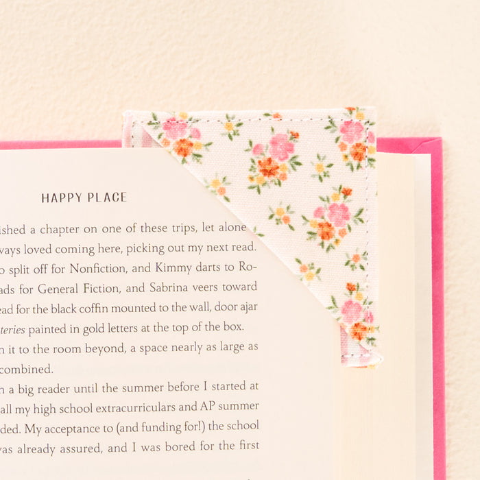 The Endless Daydream Cream Corner Keeper Fabric Bookmark by the-darling-effect, featuring a pink and green floral pattern, holds the top right corner of a book open. Charming gift for book lovers, shown on a soft, light background.
