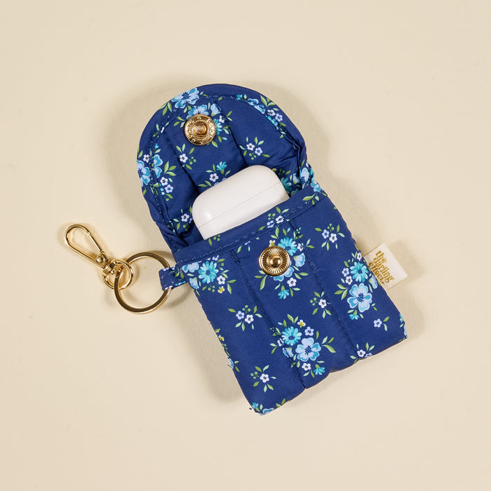 The Endless Daydream Navy Puffy Earbud Case Keychain by the-darling-effect holds a white wireless earbud charging case and features a gold snap button and keyring, shown on a beige background.