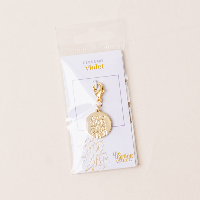 The-darling-effect's February Jewelry Birth Flower Charm Gold features a gold charm engraved with a violet flower, attached to a clasp, and displayed on a "February violet" card in a clear plastic bag for personalized gifting.