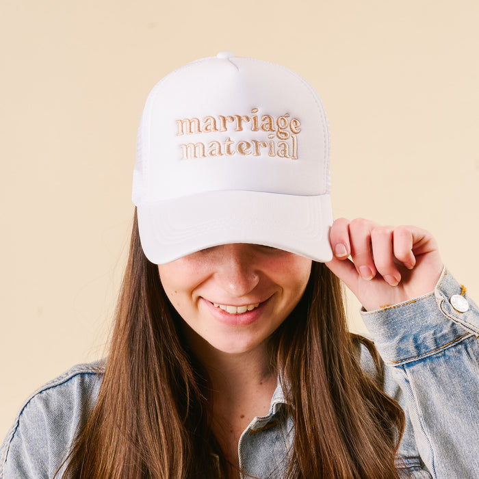 A woman with long brown hair smiles while wearing the-darling-effect's Marriage Material Trucker Hat, featuring "marriage material" embroidered on the front, paired with a light denim jacket against a beige backdrop.