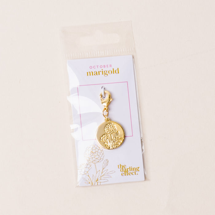 The-darling-effect’s October Jewelry Birth Flower Charm Gold features an engraved marigold on a white “October marigold” card, floral illustration, and brand logo, all packaged in a clear plastic bag.