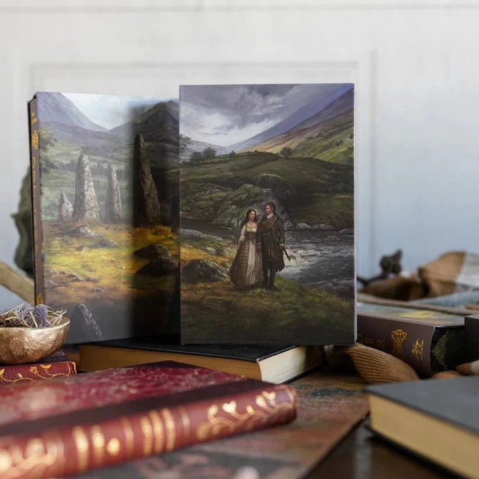 Two Outlander Annotated Special Edition slipcases propped open and displayed on vintage books, showing the illustrated slipcase exterior artwork — the Scottish Highland standing stones landscape (left) and the Jamie and Claire Fraser riverside Highland scene (right), styled with antique books, a gold bowl with dried lavender, and rustic decorative accents