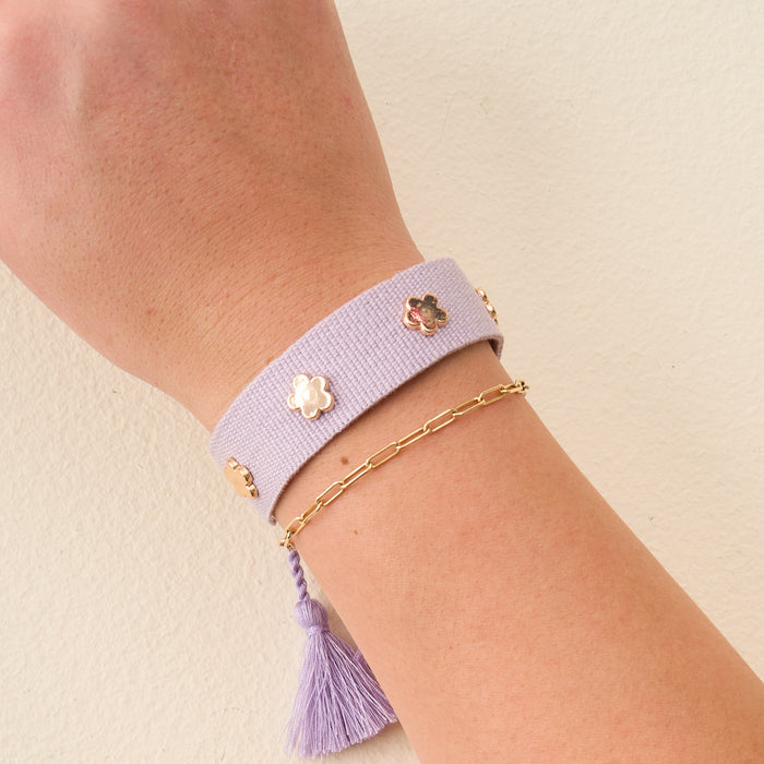 A wrist with two adjustable bracelets: The Darling Effect's Periwinkle Woven Stacker Bracelet—a wide lavender band with flower details and a purple tassel—plus a thin gold chain bracelet, shown against a light beige background.
