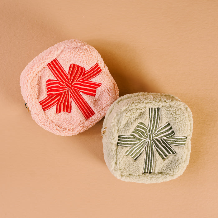 Two Square Holiday Gift Bow Red Teddy Pouches by The Darling Effect, crafted from soft sherpa—one pink with a red striped bow, the other cream with a green bow—resemble charming gift-wrapped packages on a beige surface.