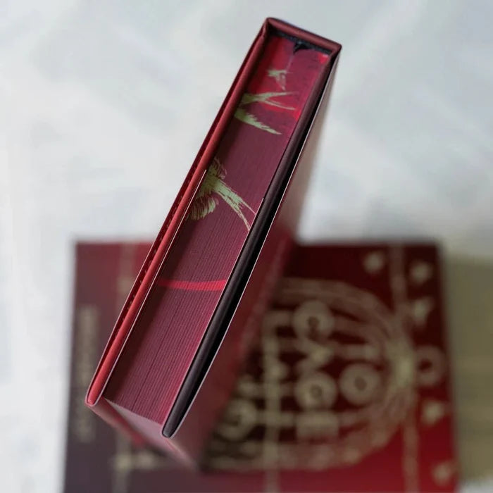 Close-up overhead view of the To Cage a Wild Bird LitJoy Edition book spine — deep red with gold foil bird and feather details and a red ribbon bookmark, resting on the gold barbed wire cage cover on a white surface