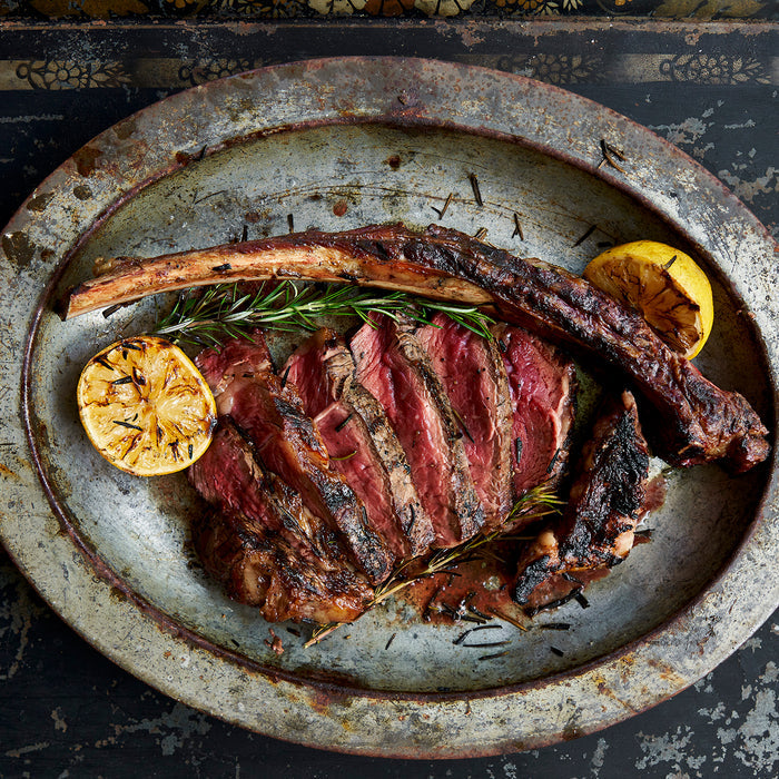 Cooked tomahawk ribeye with lemon and rosemary on metal plate. The ribeye is being served off the bone.