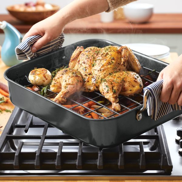 A person removes a roasted chicken from the oven, perfectly cooked in the 16.5-Inch x 13.5-Inch Nonstick Steel Roaster with Reversible Rack on the stovetop.