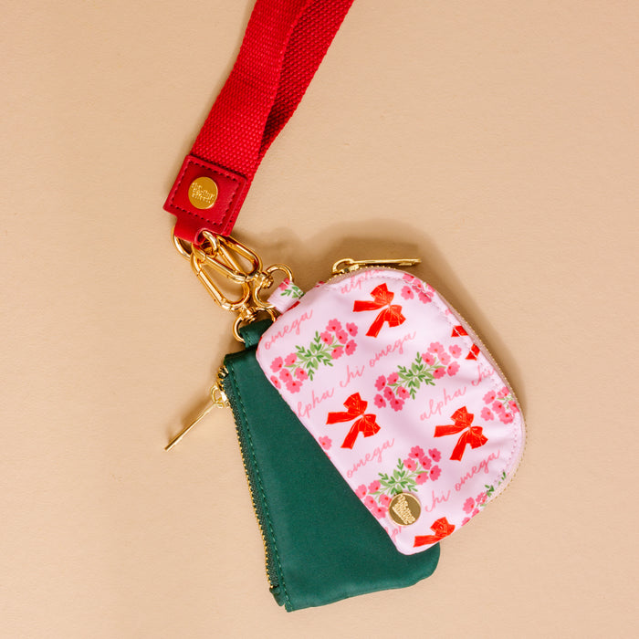 The Darling Effect Alpha Chi Omega Dynamic Duo Pouch Wristlet features two small zippered pouches—a green one and a white one with red bows, pink flowers, “aka ivy” text—attached to a red strap with gold clasps for a stylish look.