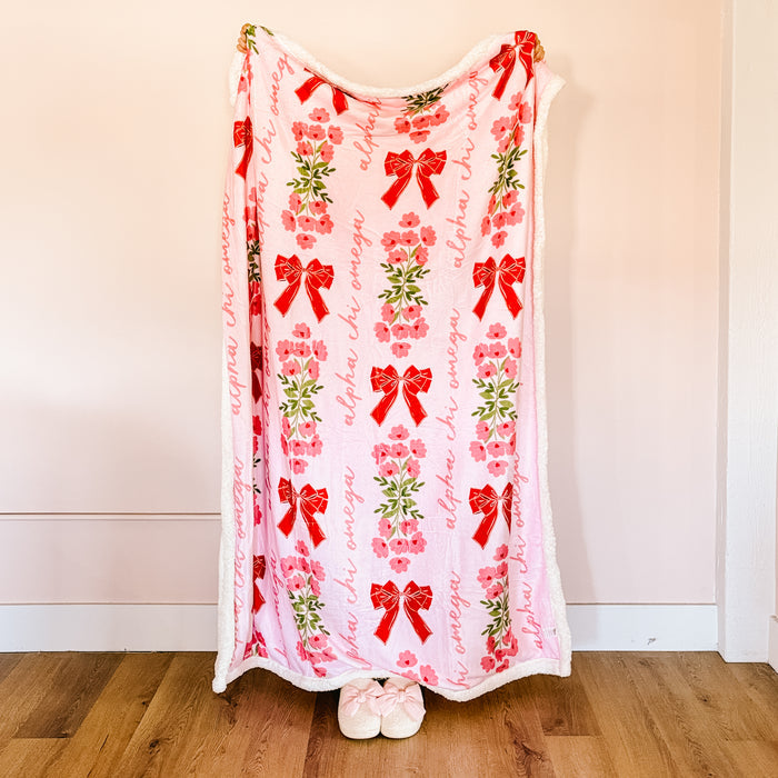 The Darling Effect's Alpha Chi Omega Plush Sherpa Blanket features soft pink sherpa, red bows, pink flowers, green leaves, and "alpha chi omega" in script. It hangs vertically against a light wall with fuzzy slippers at the bottom—a perfect sorority gift!.