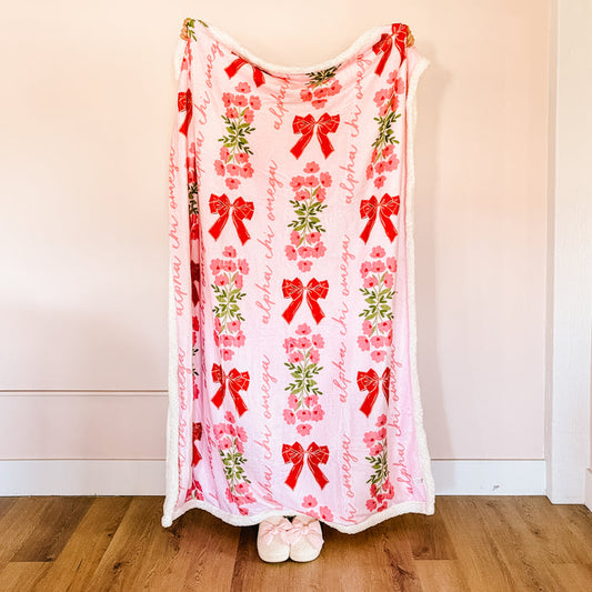 The Darling Effect's Alpha Chi Omega Plush Sherpa Blanket features soft pink sherpa, red bows, pink flowers, green leaves, and "alpha chi omega" in script. It hangs vertically against a light wall with fuzzy slippers at the bottom—a perfect sorority gift!.