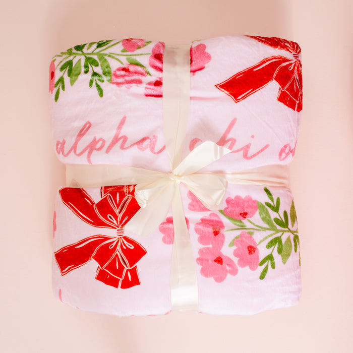 The Darling Effect’s Alpha Chi Omega Plush Sherpa Blanket features soft pink fabric with red bows and green and pink florals, neatly folded with a cream ribbon. “Alpha Chi” is partially visible on this cozy sorority blanket.