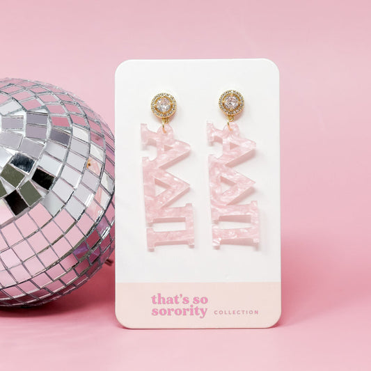 A pair of Alpha Delta Pi Letter Pink Acrylic Earrings by The Darling Effect hang on a card beside a silver disco ball—ideal for bid-day baskets or sorority events.