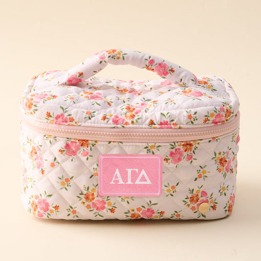 The Darling Effect Alpha Gamma Delta Glam & Go Quilted Cosmetic Bag is a floral, quilted cosmetic bag with a handle and zipper, featuring a pink patch with white “ΑΓΔ” Greek letters on the front.