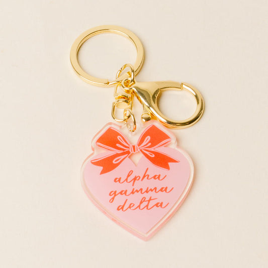 The Alpha Gamma Delta Sweetheart Bow Acrylic Keychain by The Darling Effect has a gold keychain, large clasp, pink acrylic heart charm with a red bow, and "alpha gamma delta" in cursive—an adorable gift for any sorority member.