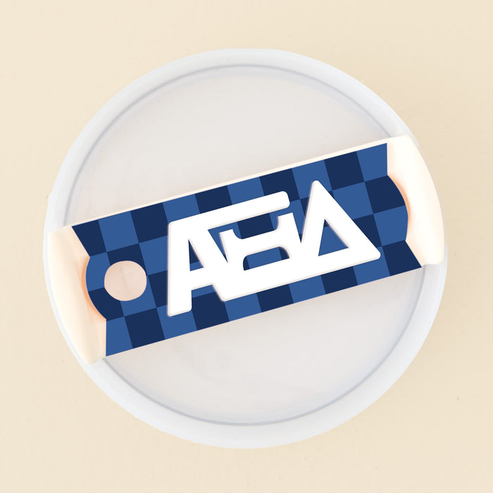 The Darling Effect's Alpha Xi Delta Tumbler Lid Tag is a round white acrylic tag with a blue and navy checkered label and bold white letters, perfect for labeling 40 oz tumbler lids or as Sorority Tumbler Lid Tags. Pale beige background.