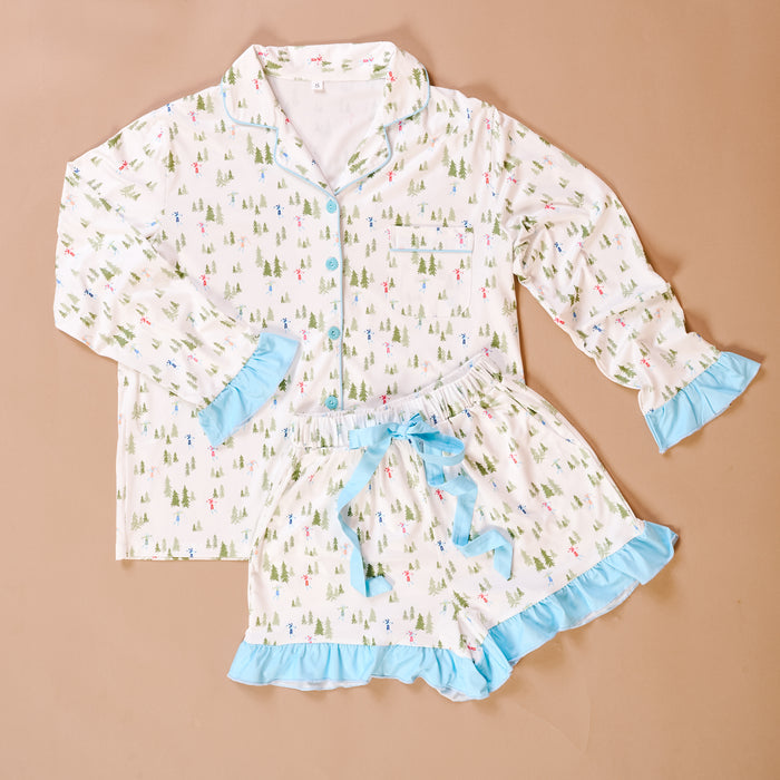 The Darling Effect's Aprés Ski Sweet Dreams Pajama Shorts Set features a white, ultra-soft two-piece with colorful tree and figure prints, blue trim, and ruffles on the cuffs and hems, displayed on a beige surface.