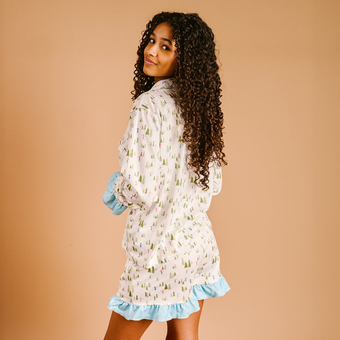 A woman with curly hair, smiling over her shoulder, models The Darling Effect Aprés Ski Sweet Dreams Pajama Shorts Set featuring a festive print and blue ruffle trim, made from ultra-soft fabric, against a plain beige background.
