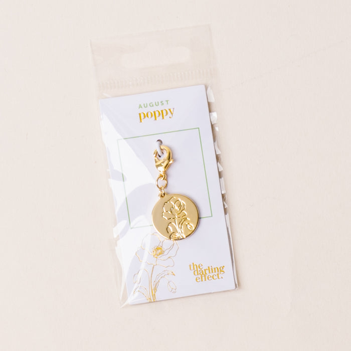 The August Jewelry Birth Flower Charm Gold by the-darling-effect features an engraved poppy flower on a gold charm with a clasp, attached to an AUGUST card, and packaged in a clear plastic bag.