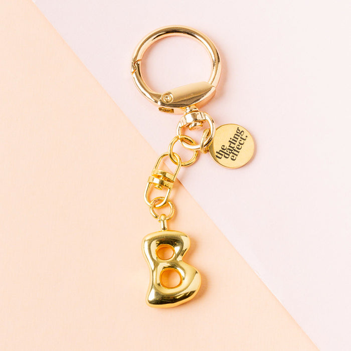 The B Bubble Letter Keychain Bag Charm by the-darling-effect features a gold bubble letter B and a round tag engraved with The Uniting Effect, set against a pastel pink and peach background.