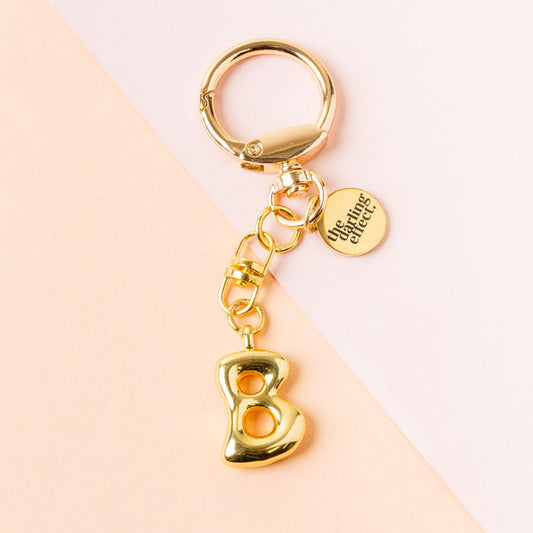 The B Bubble Letter Keychain Bag Charm by the-darling-effect features a gold bubble letter B and a round tag engraved with The Uniting Effect, set against a pastel pink and peach background.