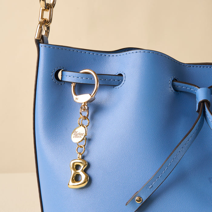 Close-up of a light blue leather handbag with gold hardware, featuring the-darling-effect’s B Bubble Letter Keychain Bag Charm—complete with a round club tag and dangling letter B—attached to the strap.