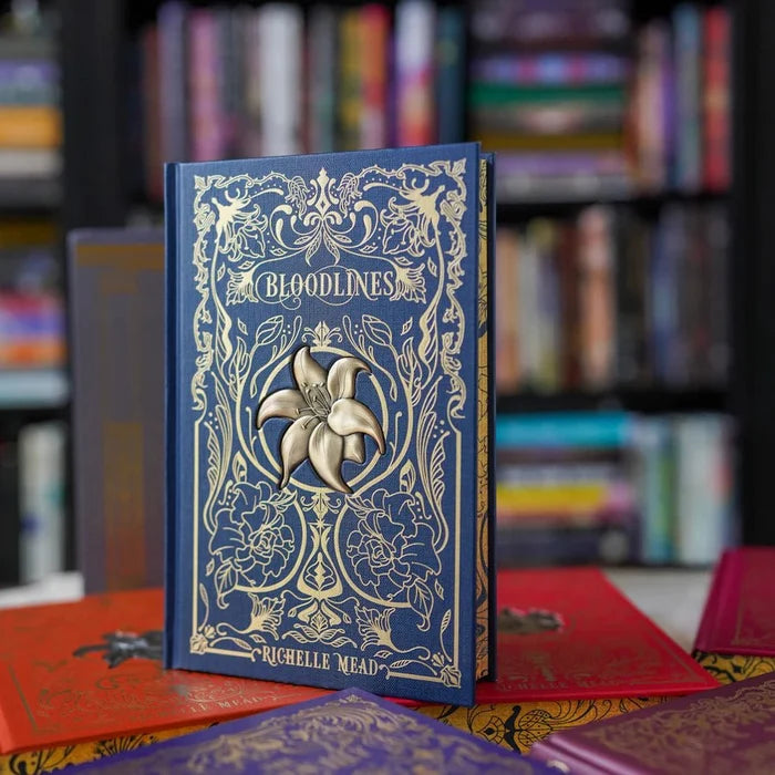 Bloodlines Special Edition Box Set by Richelle Mead – Bloodlines hardcover front cover in navy blue with gold floral design and flower ornament, standing upright with other books blurred behind