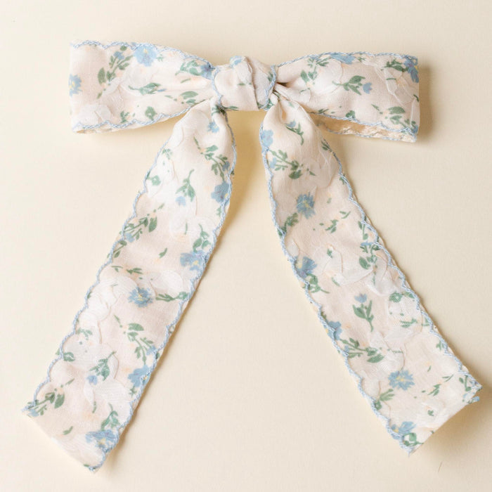 A Floral Hair Bow with a delicate floral pattern of small blue flowers and green leaves on cream fabric, displayed on a light beige surface and secured with a gold barrette clip for an elegant, embellished hair accessory.