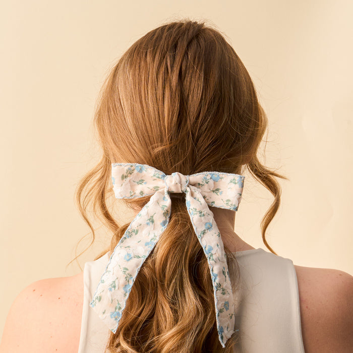 A woman with long, wavy light brown hair tied in a low ponytail with the-darling-effect’s Blue Floral Flair Hair Bow is seen from behind, wearing a sleeveless light top against a plain beige background.