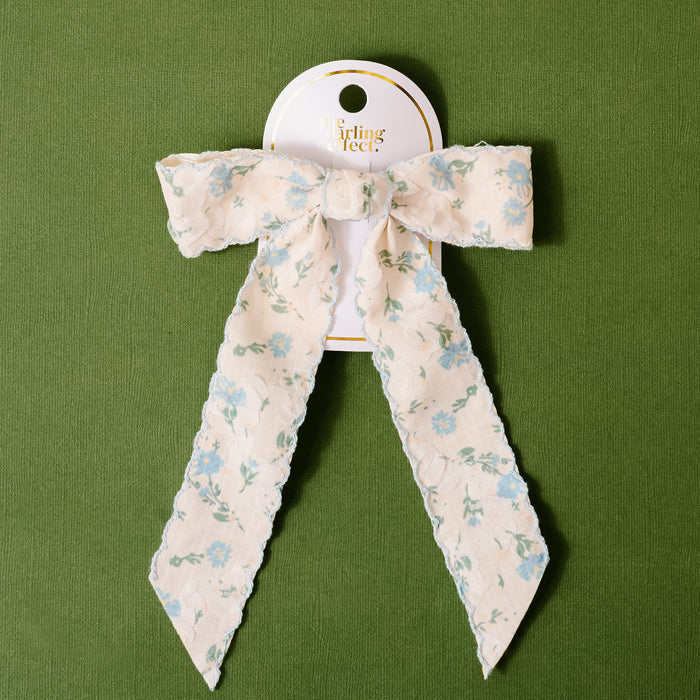 The-darling-effect Blue Floral Flair Hair Bow features blue and green flowers on white, displayed on a Girlfriends card against textured green. Includes a secure gold barrette clip for easy styling.