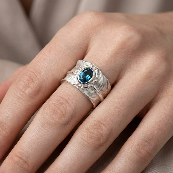 Hand wearing a textured silver feather ring with an oval blue topaz stone.
