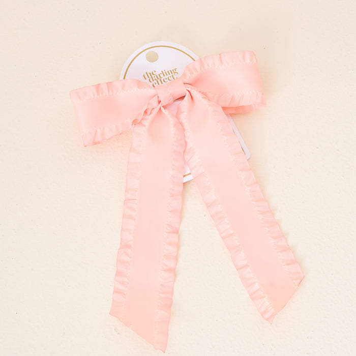 The Darling Effect’s Blush Ruffle Hair Bow features elegant ruffled satin in light pink, attached to a white tag. Ideal for bow lovers, this graceful accessory is styled simply against a soft, neutral background.