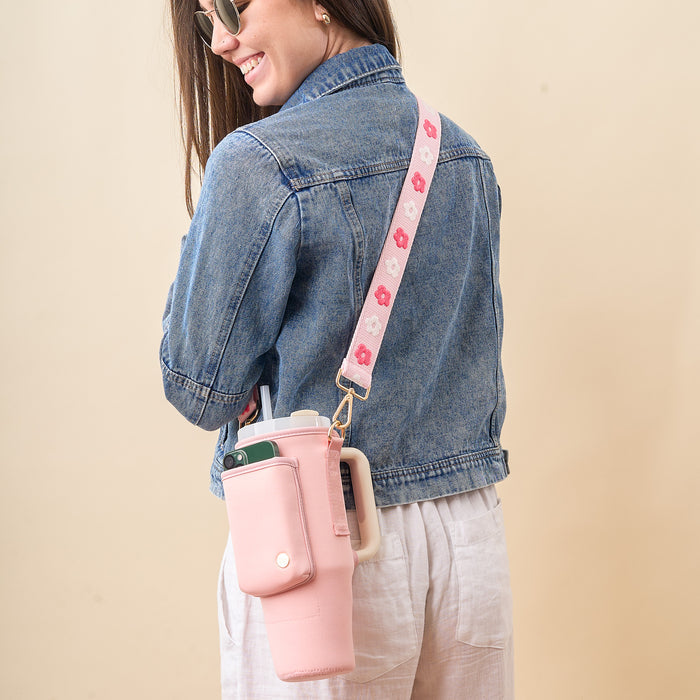 A smiling woman in sunglasses and a denim jacket carries The Darling Effect’s Blush Tumbler Sling with a floral adjustable strap and phone pocket, perfectly complementing her white pants.