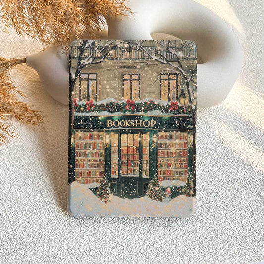 A Kindle case featuring a printed design of a bookshop with Christmas decorations, placed on a white surface with a cup and decorative items in the background.