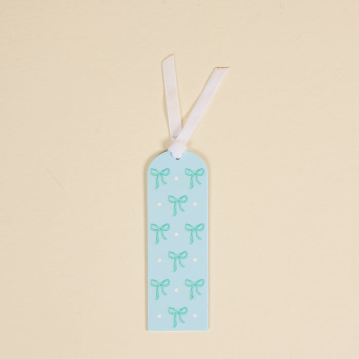 The Bows Blue Acrylic Bookmark by the-darling-effect features light teal bows and white polka dots on light blue acrylic, topped with a white ribbon—perfect for book lovers.
