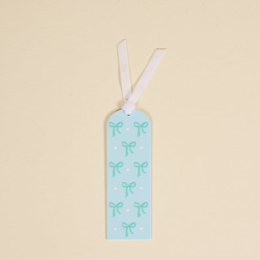 The Bows Blue Acrylic Bookmark by the-darling-effect features light teal bows and white polka dots on light blue acrylic, topped with a white ribbon—perfect for book lovers.