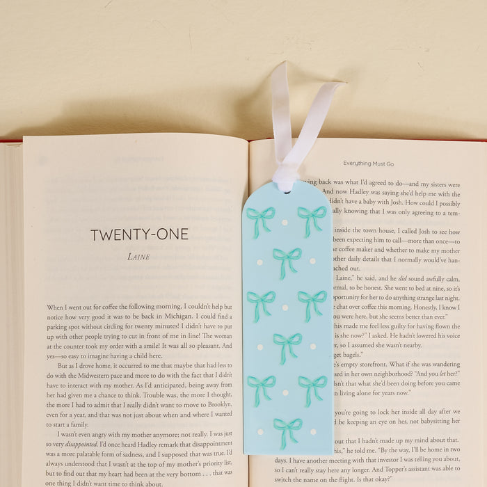 A delightful gift for book lovers, the-darling-effect’s Bows Blue Acrylic Bookmark features a green bow, white dot accents, and a white ribbon, shown atop an open book turned to chapter twenty-one on a light surface.