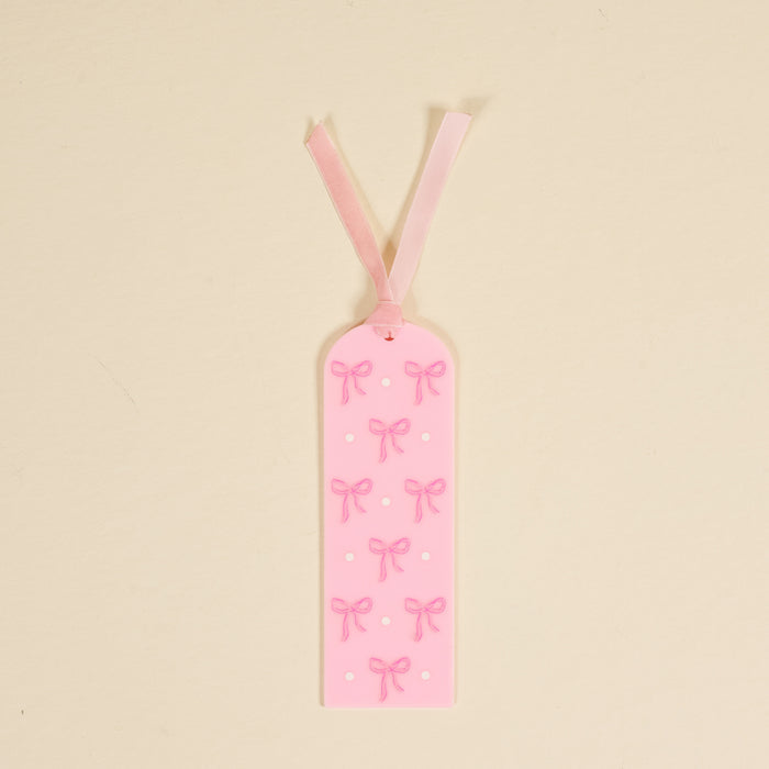 The Bows Pink Acrylic Bookmark by the-darling-effect is a perfect gift for book lovers, featuring charming bows, a pink ribbon, and white dots on a light beige background.