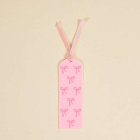 The Bows Pink Acrylic Bookmark by the-darling-effect is a perfect gift for book lovers, featuring charming bows, a pink ribbon, and white dots on a light beige background.