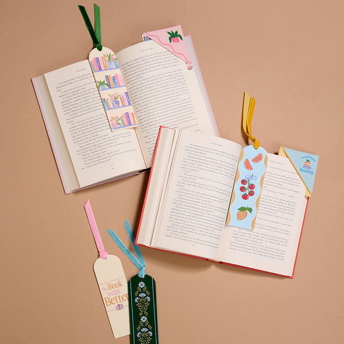 Flat lay of three open books on a tan surface, each with a Book Shelfie Acrylic Bookmark. Three additional bookmarks—with designs featuring books, plants, text, fruit, and velvet ribbon tassels—are arranged around them. Perfect for book lovers!.