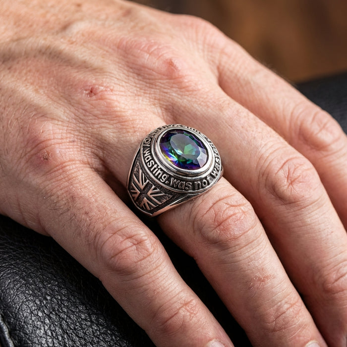 Close-up of a man's hand wearing a chunky sterling silver rock 'n' roll ring featuring a large oval mystic topaz gemstone, guitar engravings on the band, and text around the bezel, resting against a dark leather background.