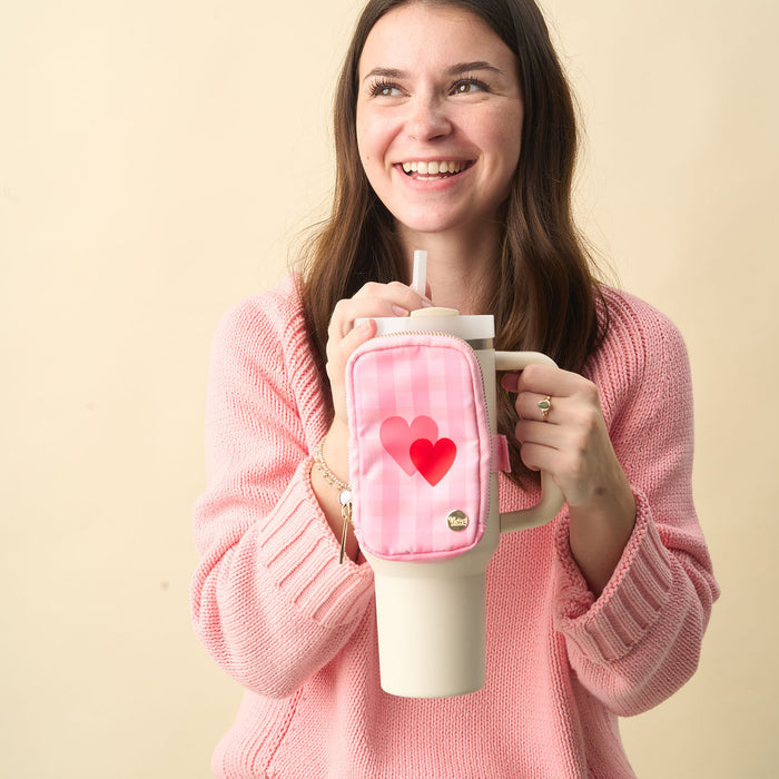 A smiling woman in a pink sweater holds a large white tumbler with a straw and The Darling Effect’s Charming Gingham Tumbler Fanny Pack, featuring two red hearts, against a plain beige background.
