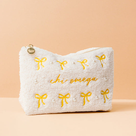 The Darling Effect Chi Omega Bows & Pearls Teddy Pouch is a fluffy white sherpa cosmetic bag with gold embroidered bows, pearl accents, and Chi Omega text—an elegant sorority gift.