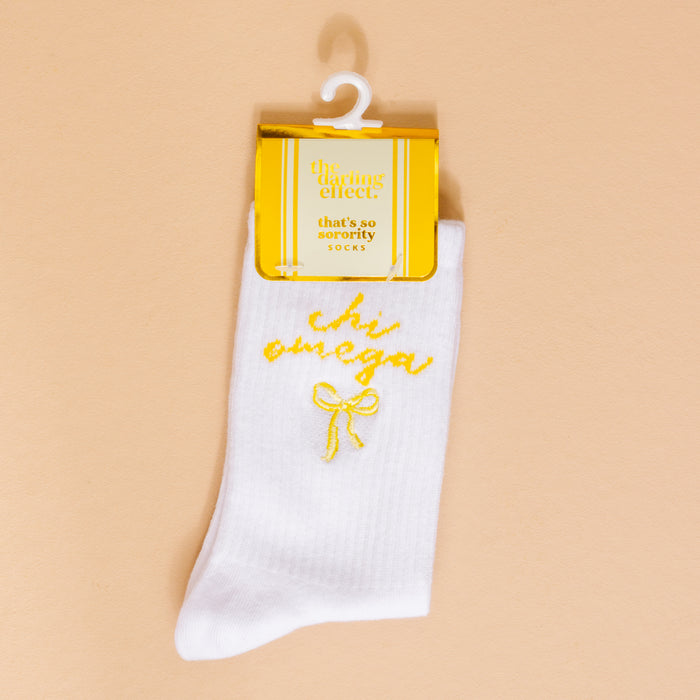Chi Omega Embroidered Crew Socks by The Darling Effect: white socks with yellow Chi Omega and bow embroidery, attached to a chic gold and cream “the darling effect” tag—cozy sorority style!.
