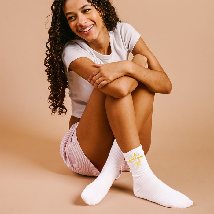 A smiling woman with long curly hair sits against a beige backdrop, wearing a white t-shirt, pink striped shorts, and The Darling Effect's Chi Omega Embroidered Crew Socks with yellow details.