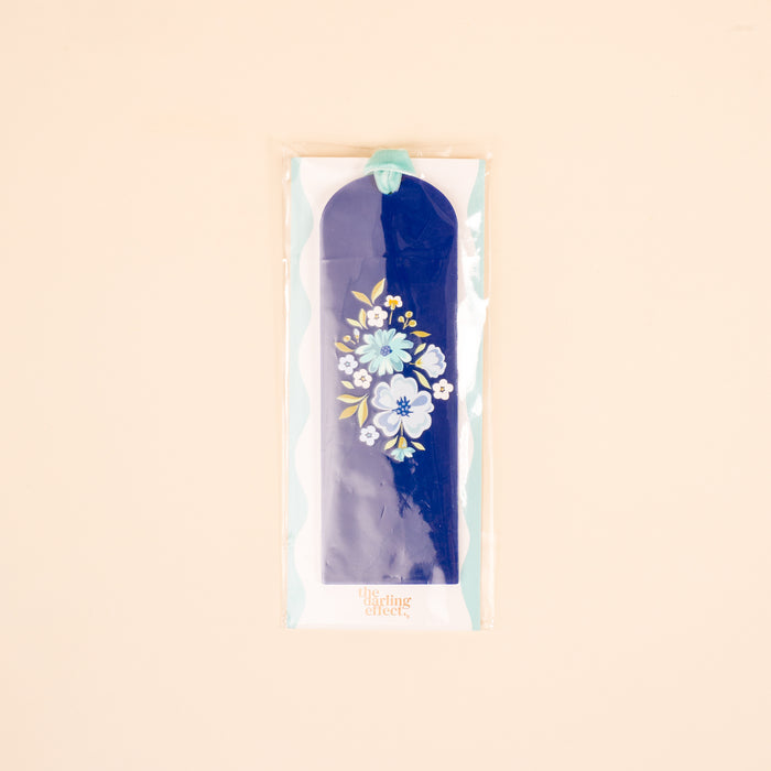The "Dainty Floral Navy Acrylic Bookmark" by the-darling-effect features a white and blue floral design, turquoise velvet ribbon tassel, and comes in a clear plastic sleeve—a perfect gift for book lovers, shown on a light beige background.