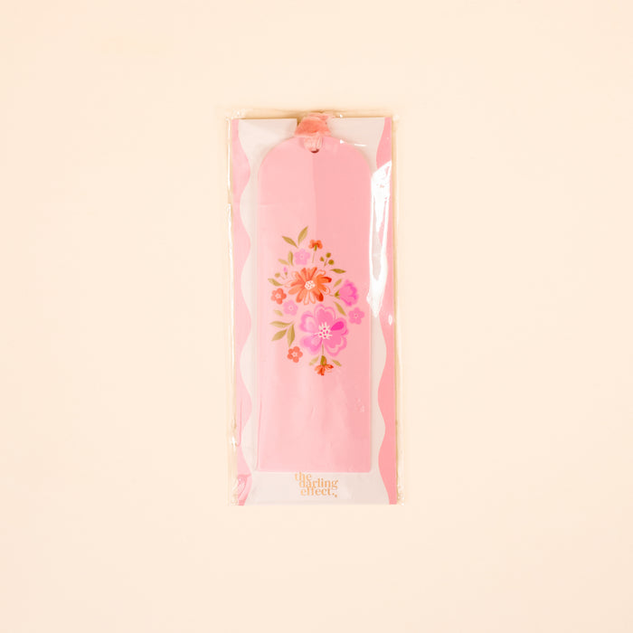 The Dainty Floral Pink Acrylic Bookmark by the-darling-effect is a lovely gift for book lovers, showcasing pink and orange flowers. It comes in a clear plastic sleeve with a pink ribbon, against a light beige background.