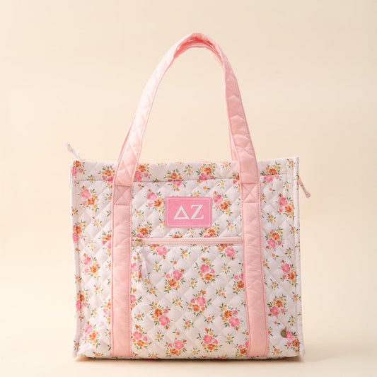 The Darling Effect’s Delta Zeta The Everyday Quilted Tote Bag features pink handles, a white quilted floral print with pink and yellow flowers, a front zip pocket, and a Greek letters patch—ideal for daily use.
